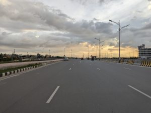 View larger photo: A four lane road with several vehicles, with a cloudy sky and beautiful orange sunlight in the background. Multiple buildings and street lights can be seen on either side of the road.