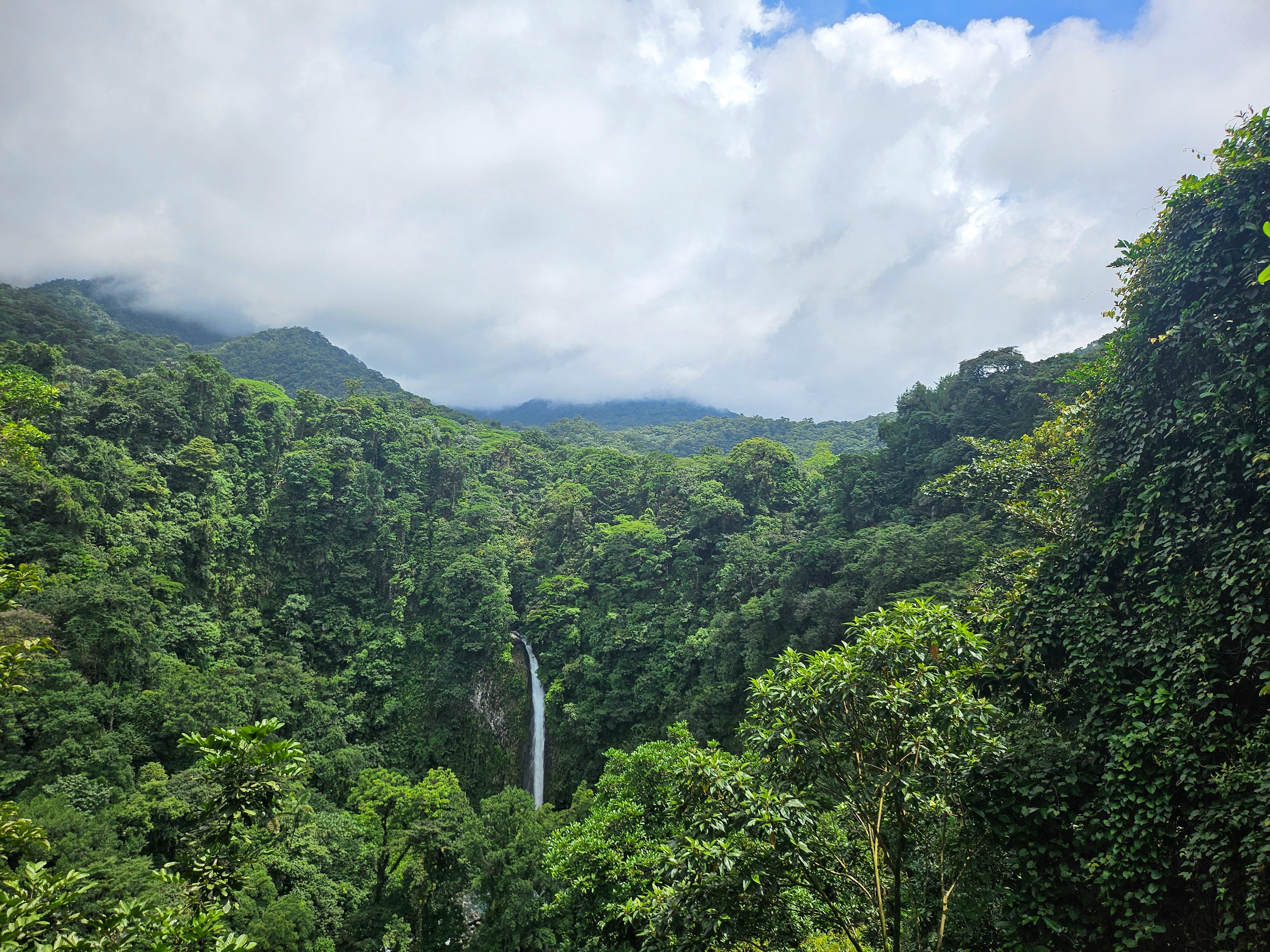 A lush green forested landscape with a tall waterfall cascading down into a hidden basin. The sky above is partly cloudy with patches of blue peeking through