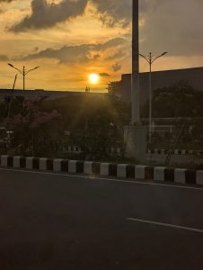 Sunset over a building. There are light poles and a road in the foreground.