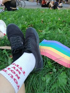 People sitting in the grass and on the road. There is a rainbow pride flag on the grass beside a person's crossed ankles and black shoes