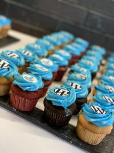 A tray of vanilla, chocolate, and red velvet cupcakes with blue frosting and WordPress or WordCamp Boston logos on top. 