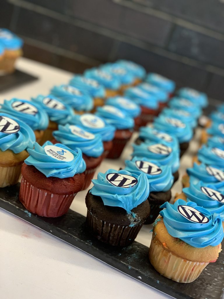 A tray of vanilla, chocolate, and red velvet cupcakes with blue frosting and WordPress or WordCamp Boston logos on top.