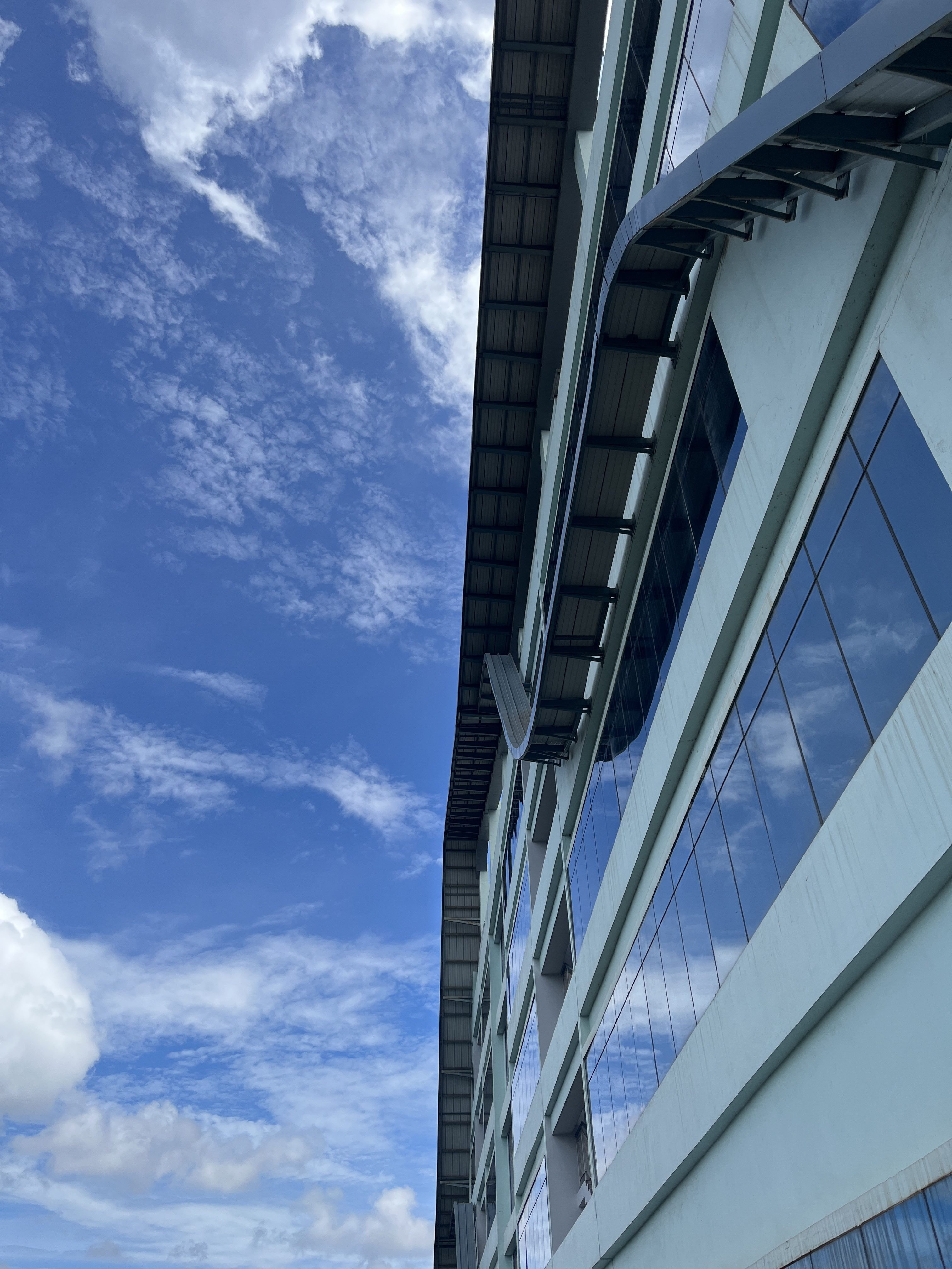 The facade of a modern concrete and glass building, seen from below, with the partially cloudy blue sky reflecting in the windows.