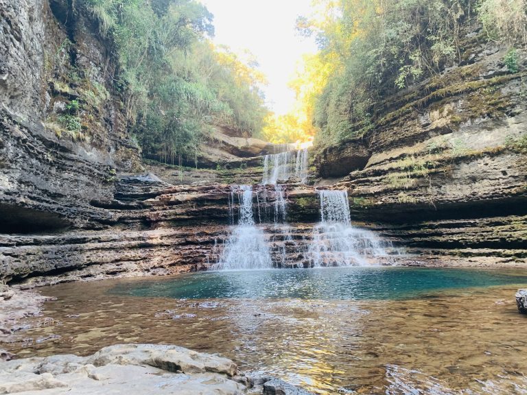 A tranquil waterfall cascades down terraced rock formations into a clear, shallow pool. The surrounding cliffs are layered with greenery and moss, with sunlight filtering through dense foliage above. The scene is framed by rugged, rocky edges and vibrant vegetation.