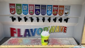  Yellow plastic cup full of shaved ice in front of ten flavors of Kona Ice’s “flavorwave” (DE Turf,  Frederica, Delaware)
