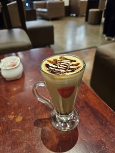 A glass of coffee with a latte art design and chocolate drizzle on top sits on a wooden table.