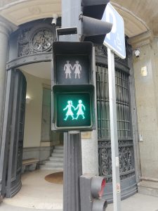 A pedestrian traffic light showing a green signal with an icon of two girls holding hands, indicating it is safe to cross. The traffic light is mounted on a post in front of a building entrance with ornate ironwork. 