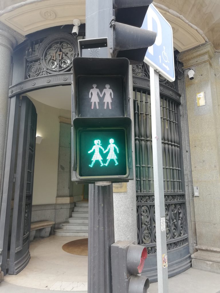 A pedestrian traffic light showing a green signal with an icon of two girls holding hands, indicating it is safe to cross. The traffic light is mounted on a post in front of a building entrance with ornate ironwork.