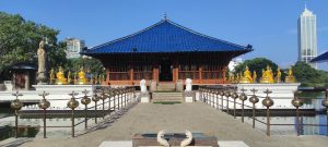 View larger photo: Seema Malaka temple. A stone bridge leads to the front door. The temple has a deep blue roof, under a blue sky.  A skyscraper can be seen in the distance.