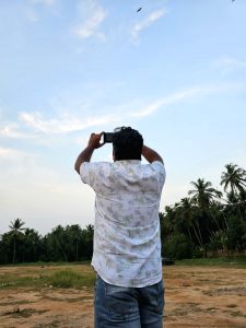 View larger photo: A man stands in a dusty field, his back to the camera, holding a smartphone up to take a picture of the birds soaring in the bright blue sky. Coconut trees line the edge of the field. 