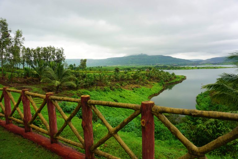 The image depicts a wooden fence overlooking a river. It is set against a backdrop of a cloudy sky and green grass. The scene captures a serene outdoor landscape during the monsoon season.