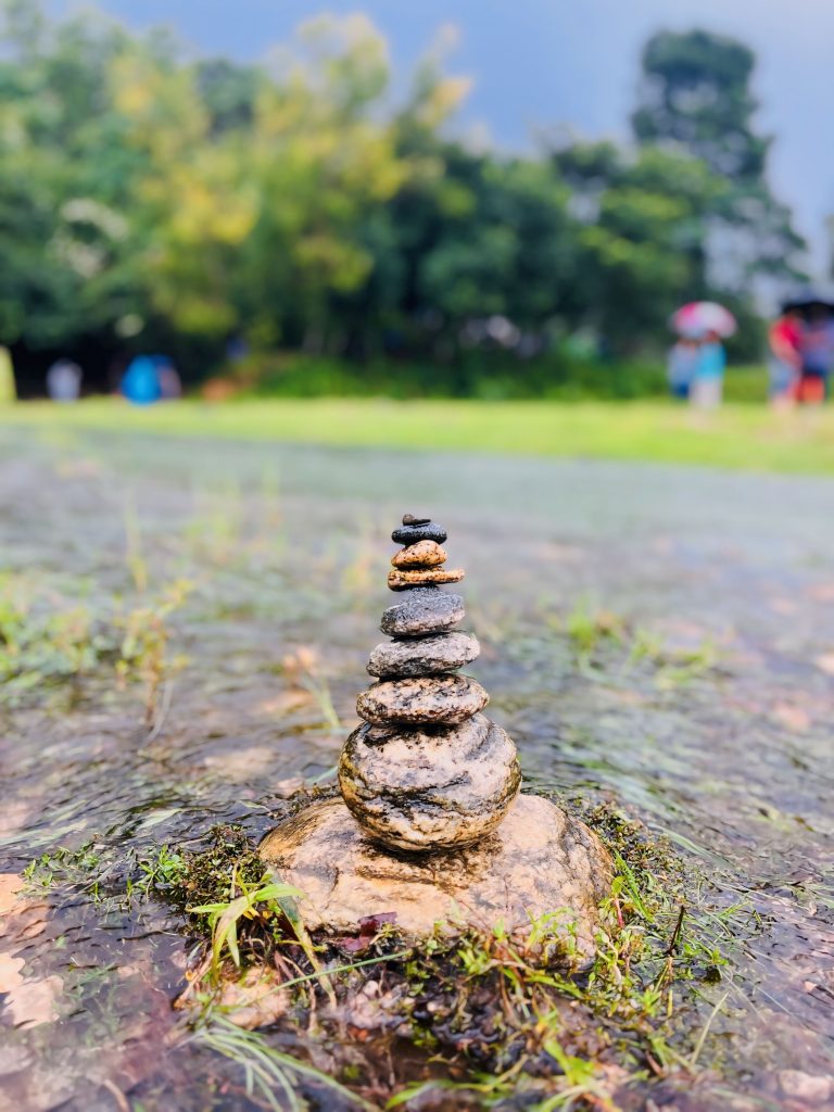 A pile of rocks in shallow water, with trees and several people in the background.