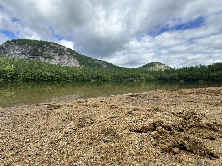 A wet, sandy beach with some footprints in the foreground with Cathedral Ledge and White Horse Ledge are visible in the distance over Echo Lake in North Conway, New Hampshire.