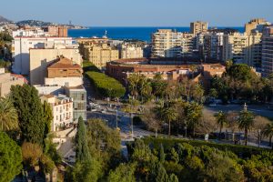 The city of Malaga taken from a hillside.  The foreground is made up of green trees and the La Malagueta bullring sits to the right of the frame set amongst modern residential tower blocks and a line of trees. The coastline and sea is visible as it forms the horizon with a deep blue sky. 