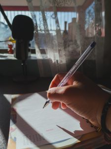 Hand writing in an agenda. Scene in front of a window with a direct ray of light on the hand and paper. A podcasting microphone and some sheer curtains can be seen in the background, out of focus.