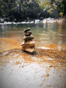 A close-up of a small stack of balanced stones on a rock in a shallow part of a flowing river. The background shows the blurred outline of the riverbank with green foliage.