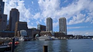 View larger photo: A waterfront cityscape with modern high-rise buildings in the background under a partly cloudy blue sky. Several boats and yachts are docked in the foreground with calm water reflecting the structures. A few seagulls are flying near the water.