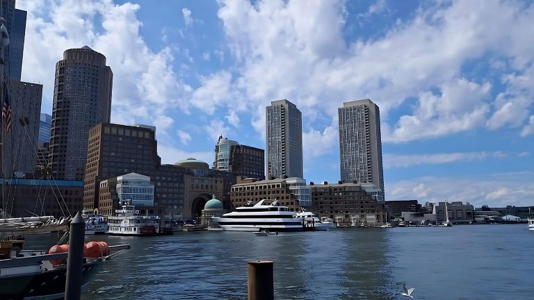 A waterfront cityscape with modern high-rise buildings in the background under a partly cloudy blue sky. Several boats and yachts are docked in the foreground with calm water reflecting the structures. A few seagulls are flying near the water.