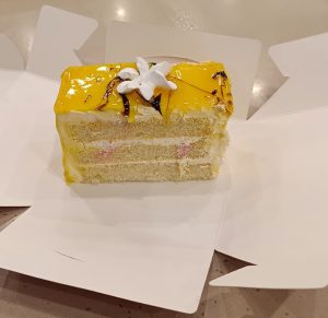 View larger photo: A piece of layered cake with yellow frosting, white cream filling, and a white star-shaped decoration on top, placed on a partially open white box.