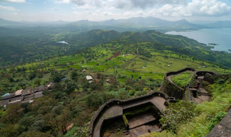 The image shows a scenic view from a hilltop or fort, overlooking a lush, green valley with fields, scattered houses, and a distant body of water.