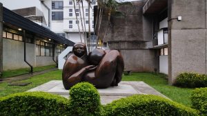 A sculpture of a human figure in a curled, seated position is displayed on a concrete base in a grassy courtyard. The background features a modern building with large windows, concrete walls, and a few palm trees. The area is surrounded by a neatly trimmed green hedge in the foreground. Located at Distance University in Costa Rica