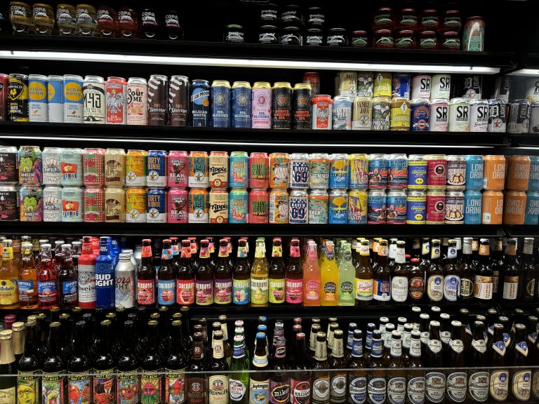 A well-stocked refrigerator display of various beers and alcoholic beverages in cans and bottles. The shelves contain a diverse selection of brands and colorful packaging, neatly organized in rows. The top shelves mostly contain cans, while the lower shelves primarily hold bottled drinks. Various flavors, styles, and types of drinks are evident from the different labels and branding.