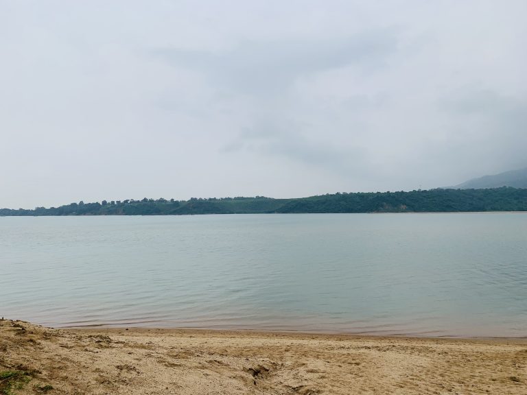 A calm, expansive body of water with a sandy shore in the foreground and a green, forested hill across the water under an overcast sky.