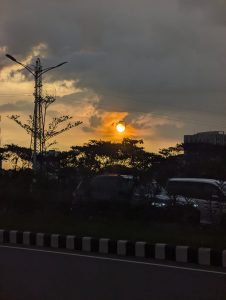 A sunset scene with the sun partially obscured by clouds, casting a golden hue over the sky.A tall streetlight and power lines are also visible on the left side of the image.