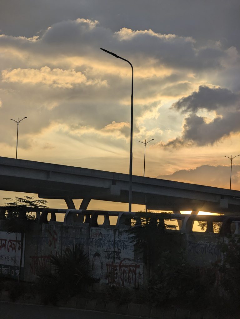 A setting sun behind a graffiti-covered concrete wall and an overpass on a cloudy evening. The silhouettes of several lightposts can be seen against the orange sky.