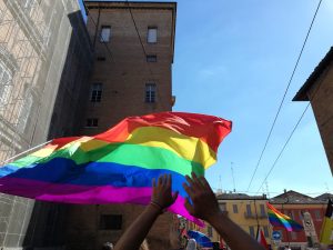 View larger photo: Hands hold up a large rainbow flag in a bright, sunny street surrounded by buildings. Other rainbow flags can be seen in the background, and the clear blue sky contrasts with the vibrant colors of the flags.