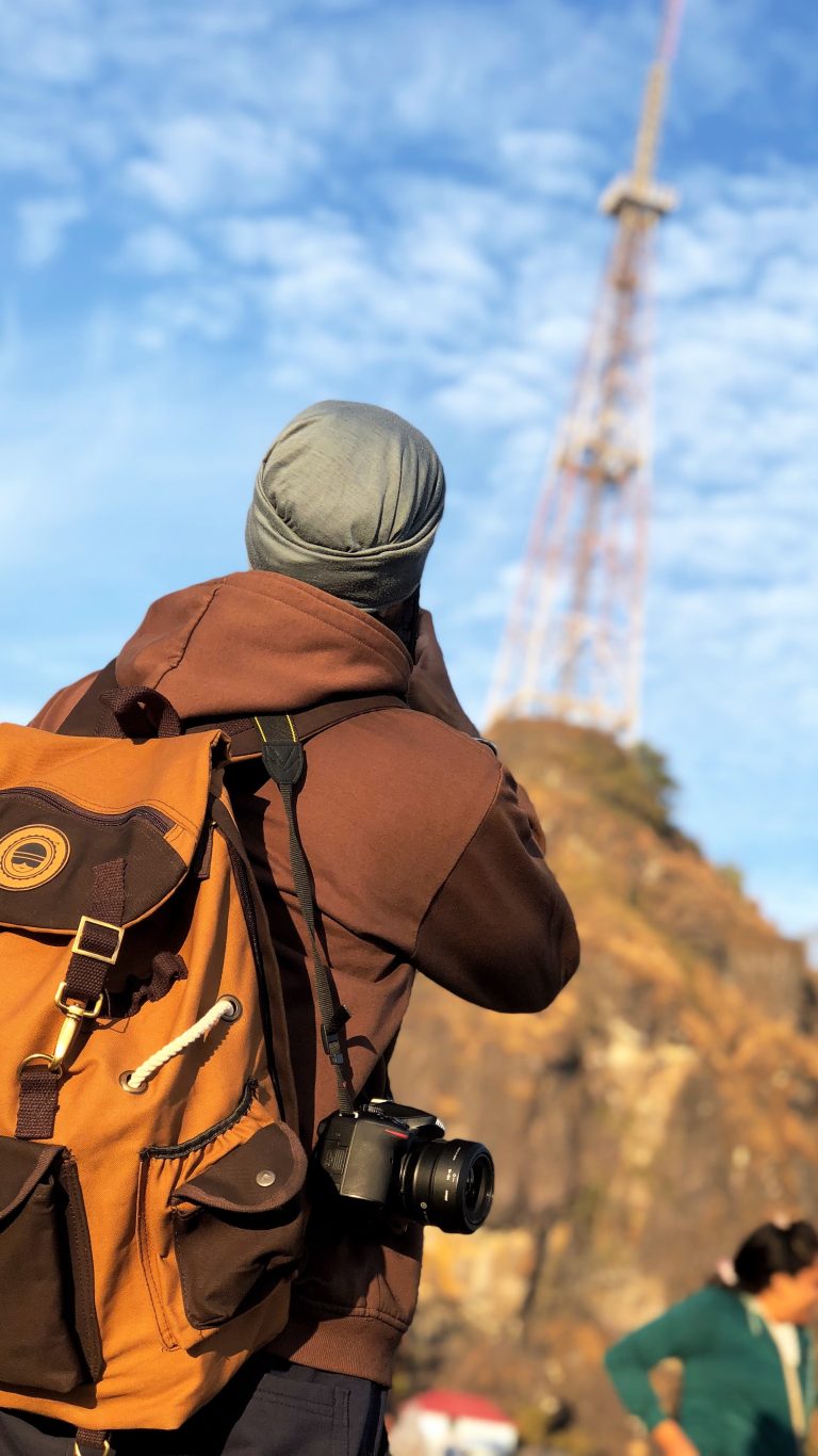 In the foreground, a person with their back to the camera looking up at a tower.  The tower in the distance in blurry whilst the person is clear. The person is wearing a jacket, a backpack, and has a nice camera.