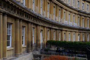 A tight crop of the corner quadrant of a crescent of regency buildings in the city of Bath in the UK.  The crescent features double pillars with ornate finishes. White railing on the balcony contrasts black railings on the street.

