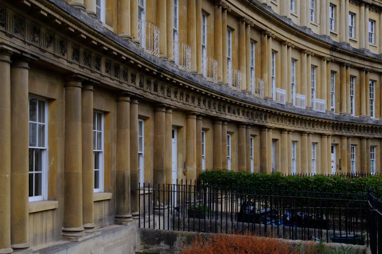 A tight crop of the corner quadrant of a crescent of regency buildings in the city of Bath in the UK.  The crescent features double pillars with ornate finishes. White railing on the balcony contrasts black railings on the street.