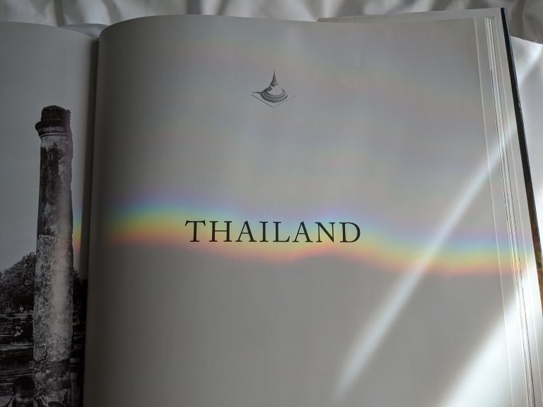 Book opened, “Thailand” on a white page with a rainbow shining over it.