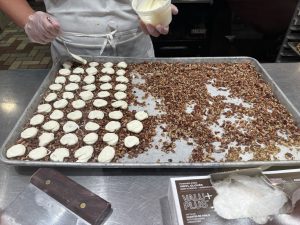 View larger photo: A person in a white apron and gloves is using a spoon to place dollops of white mixture/melted candy onto a tray filled with chopped pecans. The tray is on a metal counter and is partially covered with rows of the white mixture on top of the chopped pecans. In the foreground is a box of vinyl gloves and a metal tool with a dark handle.