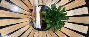 A top-down view of a restaurant table with a radial wooden pattern, featuring a potted plant and a wooden organizer holding menus, sauces and napkins.