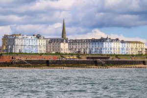 A view from the sea of an English seaside town. The sea meets a sandy beach about a third of the way up the frame with a group of buildings of pastel colours, in the centre a church spire points to a cloudy sky. 