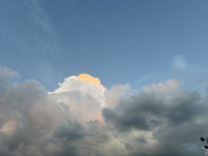 A sky filled with large, thick cumulus clouds in shades of white, gray, and hints of orange, indicating the light of the setting or rising sun. The sky above the clouds is mostly clear and blue with some faint wisps of cirrus clouds.