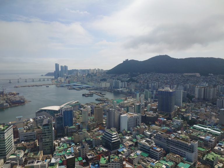 The densely built city of Busan from above, with sea and mountain in the background.