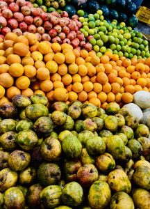 View larger photo: A vibrant display of various fruits neatly stacked in a market. The image shows an abundance of oranges, pomegranates, mangoes, melons, and green limes, arranged in separate, colorful sections