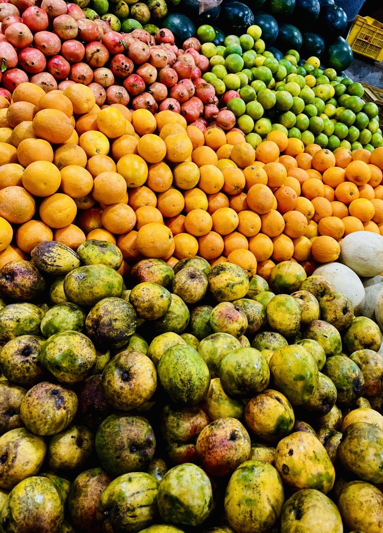 A vibrant display of various fruits neatly stacked in a market. The image shows an abundance of oranges, pomegranates, mangoes, melons, and green limes, arranged in separate, colorful sections