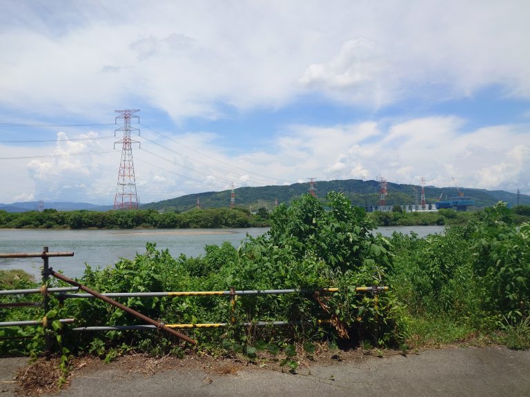 Old rusty railing along a road, overgrown with greenery. Beyond, a river, and on the other side, power lines going over water and into the mountains.