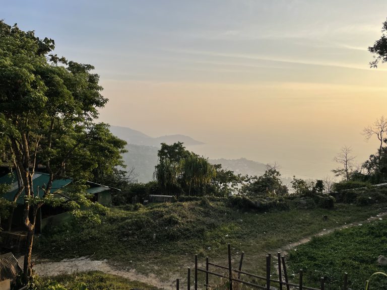 A peaceful sunset view over misty hills and the sea, with trees and a dirt path in the foreground.