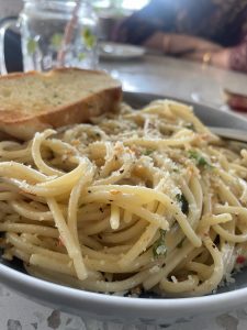 View larger photo: Close-up photograph of white spaghetti with sprinkled cheese, herbs and garlic bread. The folded arms of a person on the other side of the table can be seen in the background.