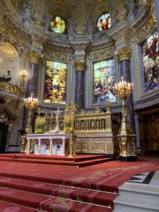 The steps to the alter inside the Berlin Cathedral. There’s red carpet on the stone steps, candles lit on either side of the alter, gold trim, and detailed religious paintings on the windows. 