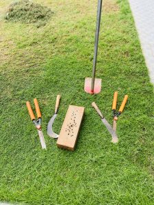 A collection of gardening tools, including pruning shears, a sickle, and a knuckle knife, placed on the grass beside a wooden block and a pole with a red blade. In the background, there is a pile of cut grass.