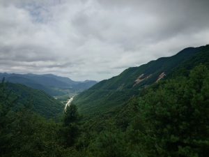 There is a big mountain with green trees and bushes on the right, with clouds and more green mountains in the background. Part of the road to the valley is visible.