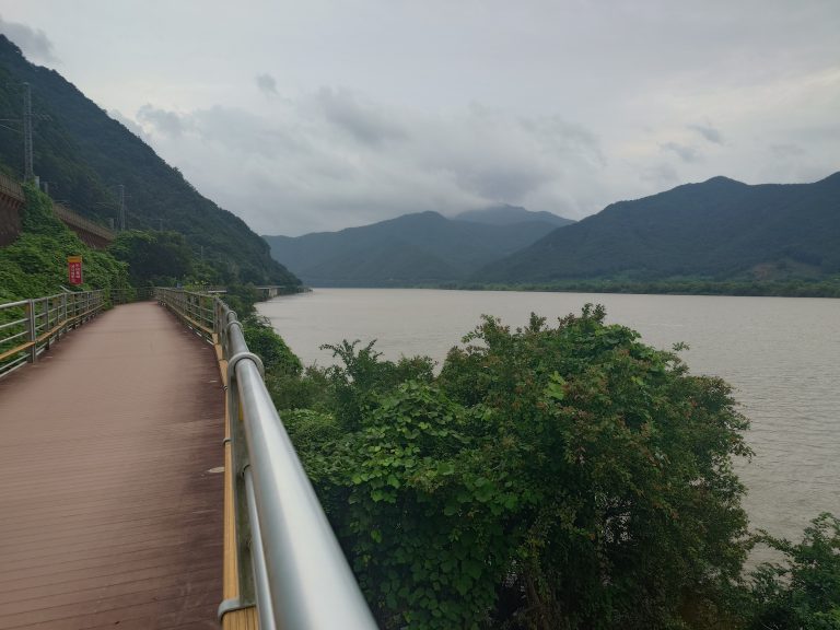 Brown bike path alongside river, some bushes, mountains on the background.