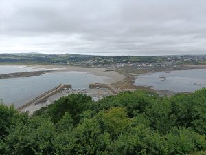 View larger photo: The view from St Michaels Mount towards Marazion. A beach on a cloudy day, the tide is out and the sea is separated by a narrow causeway that people are walking along. In the foreground the tide is out at the harbour and the boats are beached.