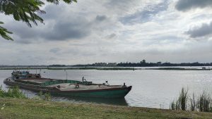 View larger photo: Peaceful river bank scene featuring a boat tied up on the shore, under a cloudy sky.